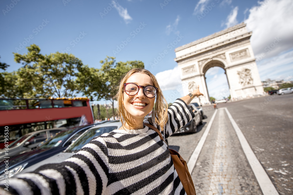 Fototapeta premium Młoda kobieta turystyczny robi selfie portret z sławnym Triumfalnym łukiem na tle w Paryż
