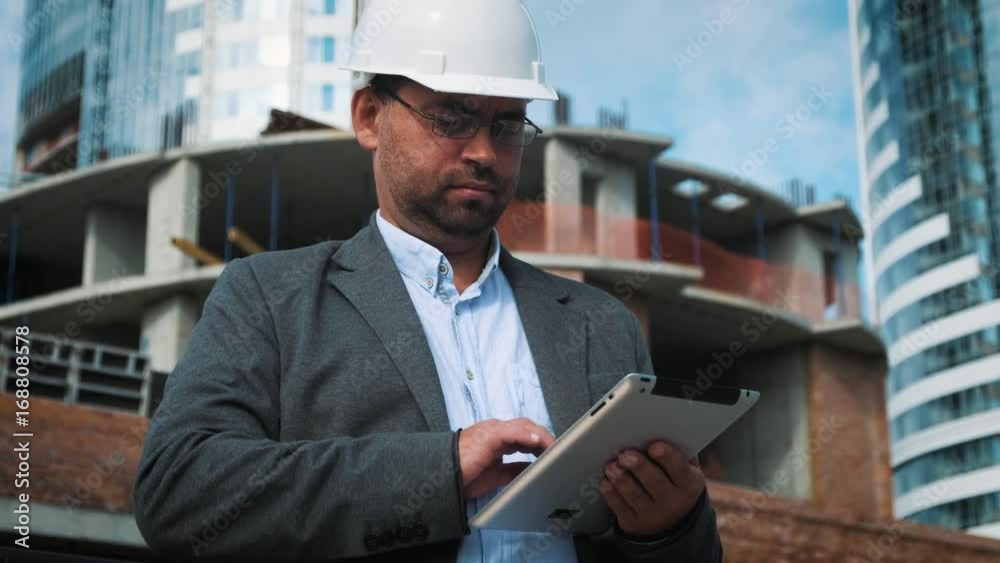 Adult engineer or architect uses a tablet in operation. Writes a message or checks a drawing. Against background is building. Builders are building a modern residential building of glass and concrete. Adult engineer or architect uses a tablet in operation. Writes a message or checks a drawing. Against background is building. Builders are building a modern residential building of glass and concrete.