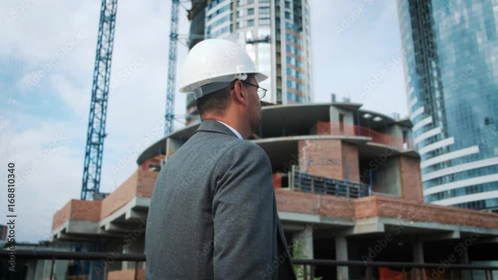 The engineer or the builder checks and controls the object under construction. He goes by the construction site, the workers are working. Against the background of innovative modern buildings. The engineer or the builder checks and controls the object under construction. He goes by the construction site, the workers are working. Against the background of innovative modern buildings.