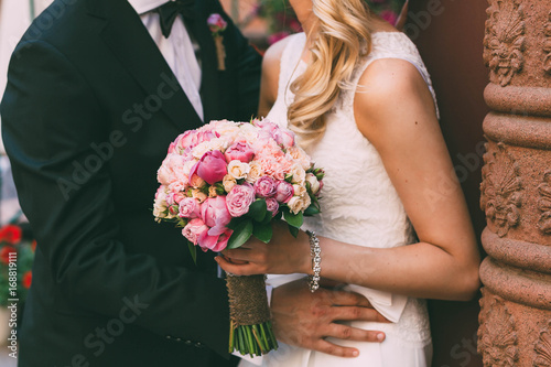 Beautiful tanned bride in a white fitting dress and groom in tuxedo are standing next to the wall and holding a wedding bouquet. Groom embrace woman by the waist. Outdoors