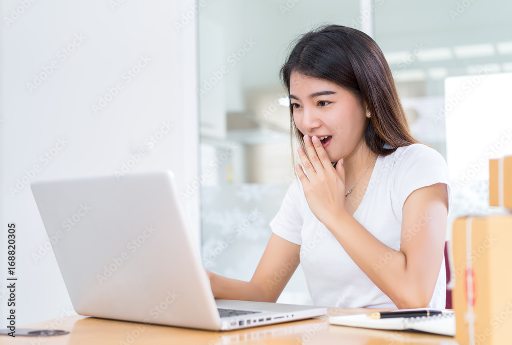 Young asian woman business shocked reaction summer sale fashion online surprise looking at laptop computer sitting on working wooden table at home office with note.