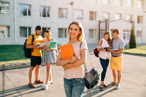 Student. Enjoying university life. Handsome young girl with grey velvet backpack holding books and smiling while standing against university with her friends in the background
