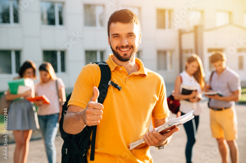 Young attractive smiling student showing thumb up outdoors on campus at the university. Selective focus photo