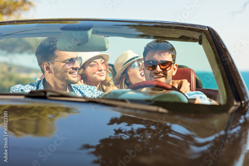 Group of cheerful young friends driving car and smiling in summer