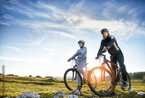 Fototapeta Naklejka Na Ścianę i Meble -  Cyclist couple with mountain bikes standing on the hill under the evening sky and enjoying bright sun at the sunset.