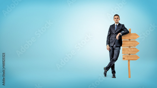 A businessman on blue background casually leans on a small wooden signboard showing way in only one direction.