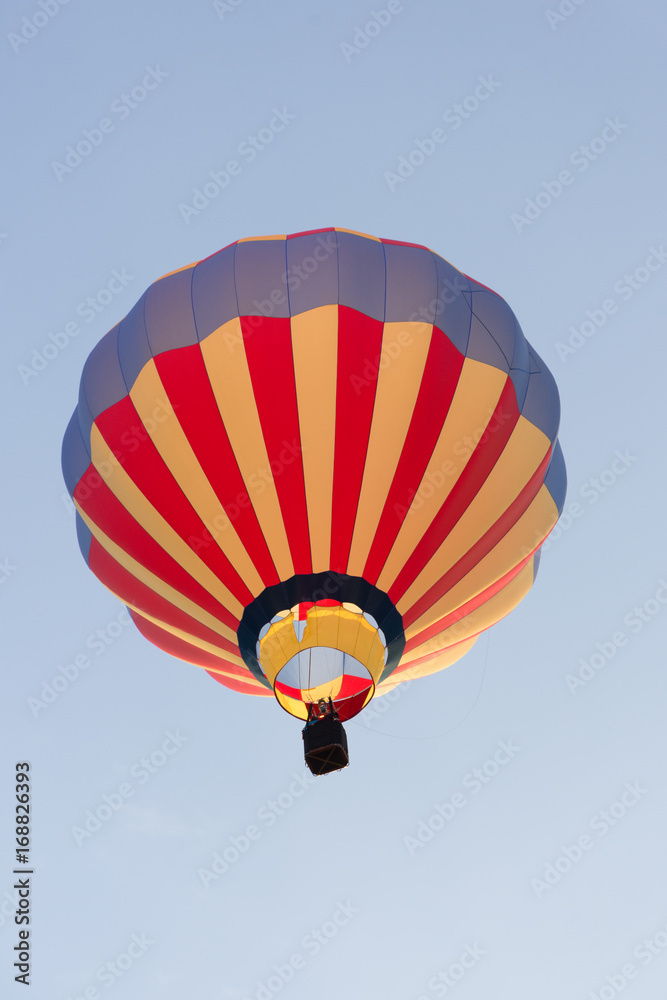 Fototapeta premium A hot air balloon in flight with red and gold vertical stripes and a blue top. The wicker basket carrying passengers is photographed from below. Copy space is available in the light blue sky.