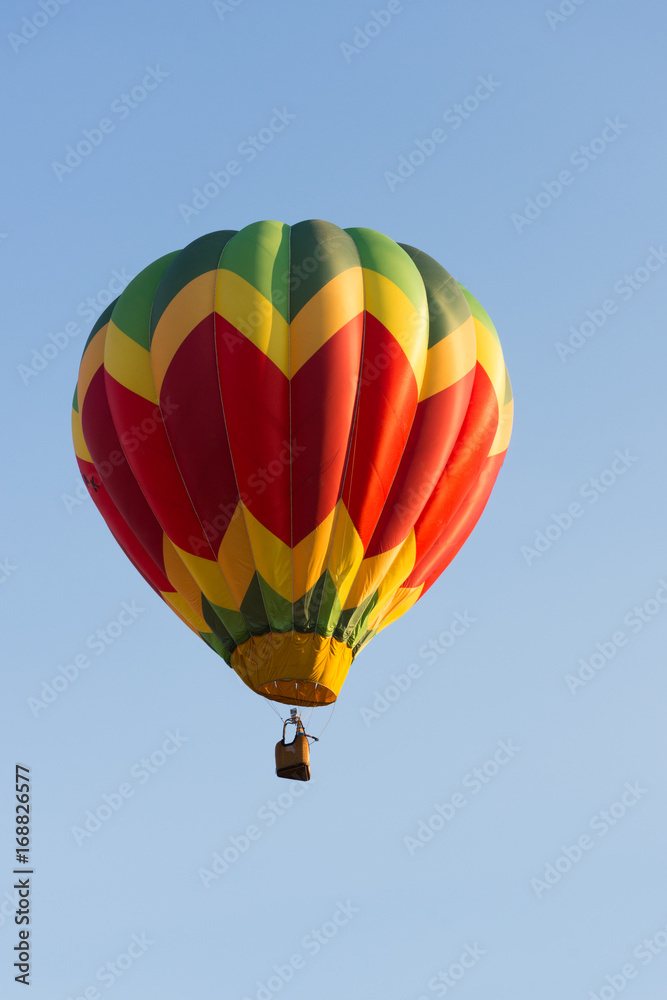 Naklejka premium Yellow, Red and Green Balloon in Flight with cloudless sky behind. The balloon colors make a chevron design. The wicker basket carries passengers that are not recognizable.