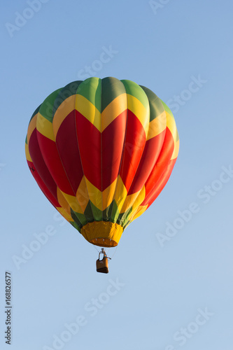 Yellow, Red and Green Balloon in Flight with cloudless sky behind. The balloon colors make a chevron design. The wicker basket carries passengers that are not recognizable. 