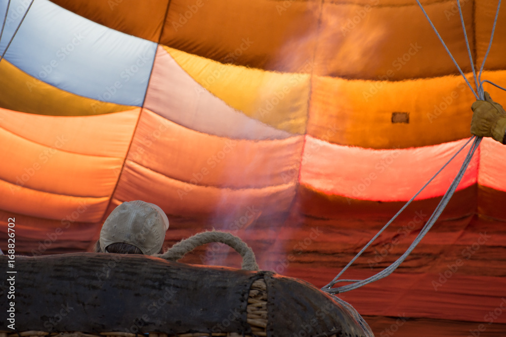 Obraz premium Close up of the flame directed by the pilot into a hot air balloon to inflate it. The balloon is stabilized by ropes a second person is holding. The colorful fabric backlit by the sun.