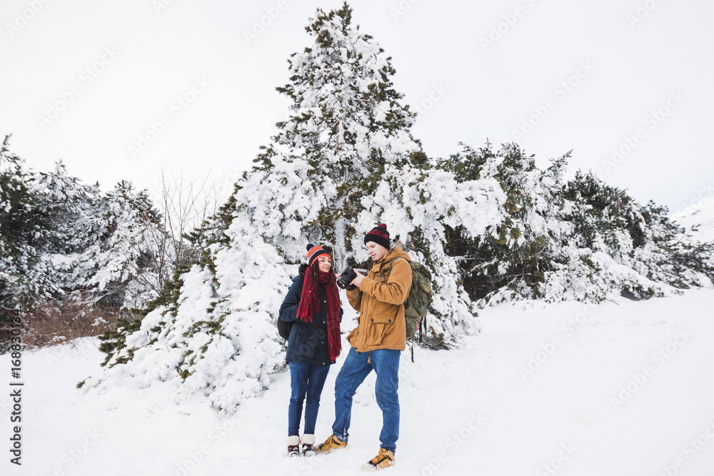Couple tourists in winter forest. Casual style, beige parka, jeans, red scarf. Taking photo of landscape Couple tourists in winter forest. Casual style, beige parka, jeans, red scarf. Taking photo of landscape