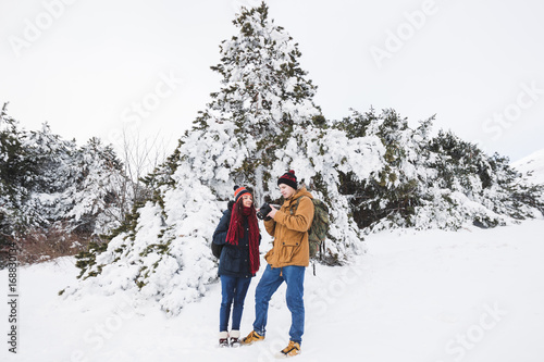 Couple tourists in winter forest. Casual style, beige parka, jeans, red scarf. Taking photo of landscape