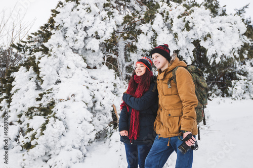 Couple tourists in winter forest. Casual style, beige parka, jeans, red scarf. Taking photo of landscape
