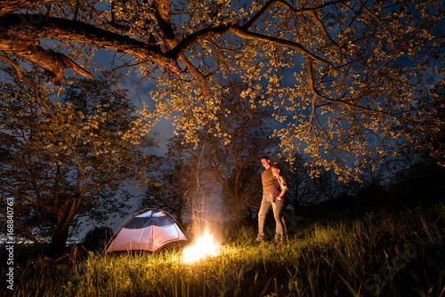 Romantic couple tourists standing at a campfire near tent  hugging each other under trees and night sky. Night camping