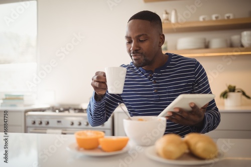 Man using a digital tablet while having cup of coffee in kitchen