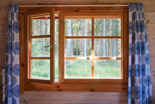 Open window with natural wooden frame and blue flower curtains. Morning forest view. Sunrise in the country