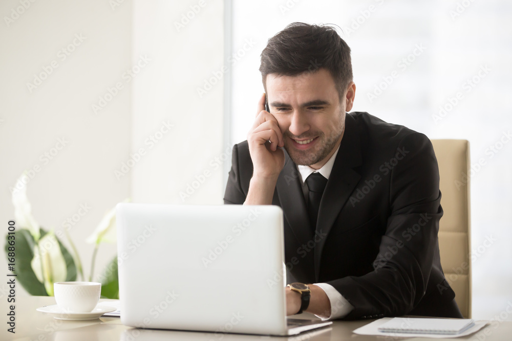 Smiling businessman talking on cellphone sitting at office desk in front of laptop, having successful mobile negotiations with client, manager consulting customer by phone, easy internet banking Smiling businessman talking on cellphone sitting at office desk in front of laptop, having successful mobile negotiations with client, manager consulting customer by phone, easy internet banking