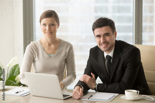 Smiling businessman and businesswoman looking at camera, confident successful boss and professional assistant posing at workplace sit in front of laptop, talented motivated business team portrait