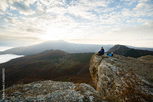 Smiling man hiker with backpack siting and relaxing on the top of the mountain and looking at beautiful yellow autumn landscape sunset over clouds