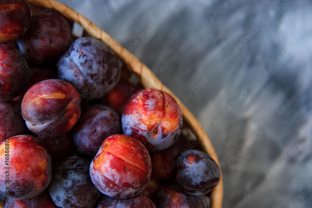 Fototapeta premium Overhead still life of purple plums in basket on grey textured background with copy space