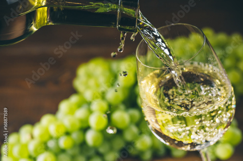 Pouring white wine into a glass with a bunch of green grapes against wooden background photo