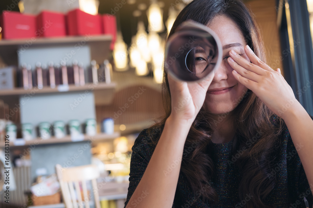 A beautiful Asian woman roll a book and looking through it with one eye while closing another eye with feeling happy , sitting in cafe A beautiful Asian woman roll a book and looking through it with one eye while closing another eye with feeling happy , sitting in cafe
