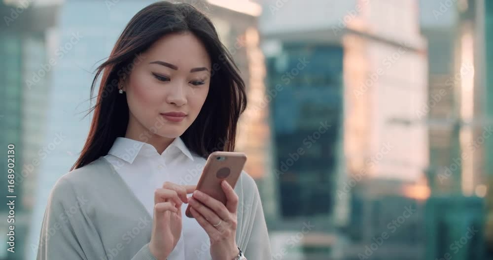 beautiful young business Asian woman in a good mood with smartphone texting messenger with skyscrapers in the background