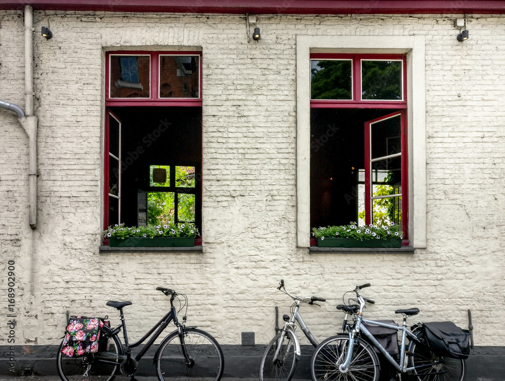 Cute bikes parked against the white bricked wall of a tavern with open windows, red frames, flowers in them. Location: Belgium