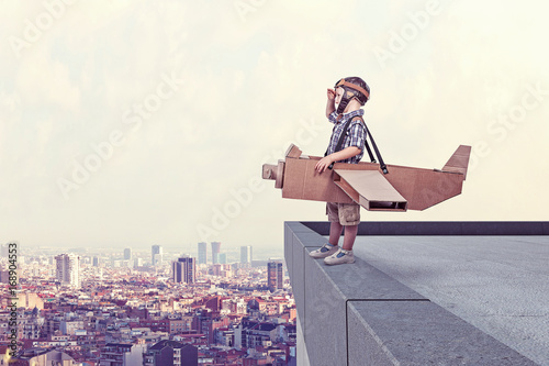  boy  cardboard airplane model  pilot dress looks away ledge  high building. city ​​in the background.  dream and imagination.