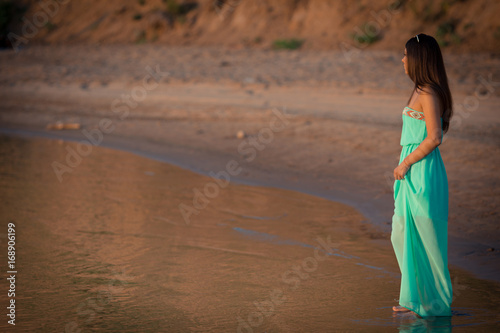 A young woman in a long dress is standing barefoot on the sand near the river