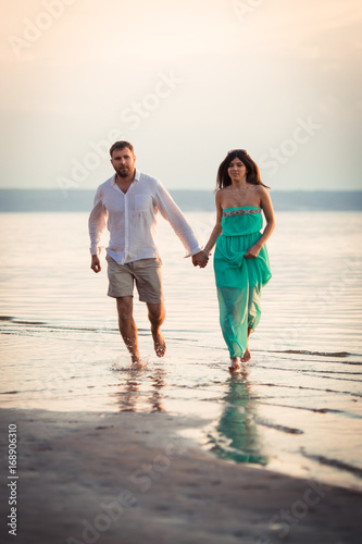 Husband and sweet wife unhurriedly run along the riverbank barefoot, their feet are wet, maybe the couple celebrate the date, the woman supports the dress