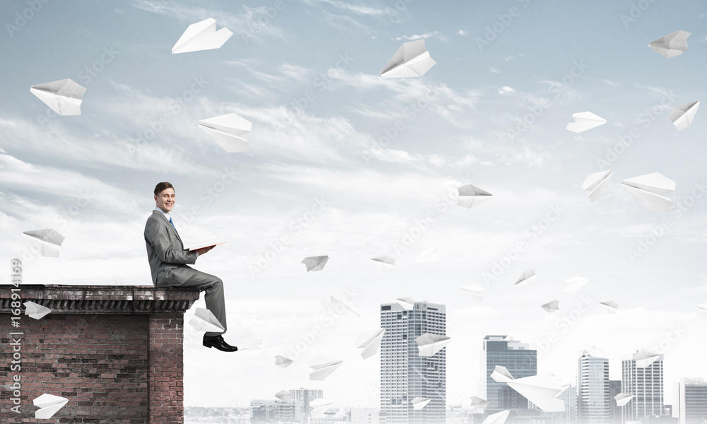 Man on roof edge reading book and cityscape at background Man on roof edge reading book and cityscape at background