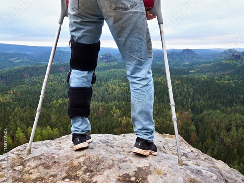 Man with leg in knee cages and crutches for stabilization stay on rock. Hurt tourist walk in mountains
