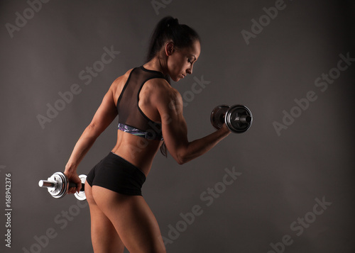 Muscular beautiful fitness model woman athlete standing posing in sportswear top and shorts, doing workout with high weight dumbbells. Copy space. Healthy life bodybuilding lifestyle concept image.