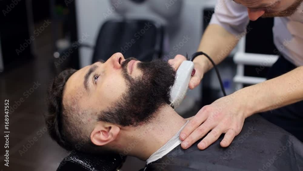 Male Barber shaves the beard of a brutal man using an electric shaver in a modern barbershop. Close-up. Male Barber shaves the beard of a brutal man using an electric shaver in a modern barbershop. Close-up.