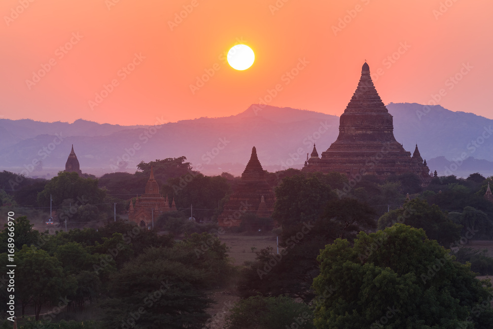 View from afar of the ancient pagodas (stupas) visible among rugged fields and trees of other pagodas and mountains on the horizon during sunset or sunrise, in Bagan, Myanmar (Burma)