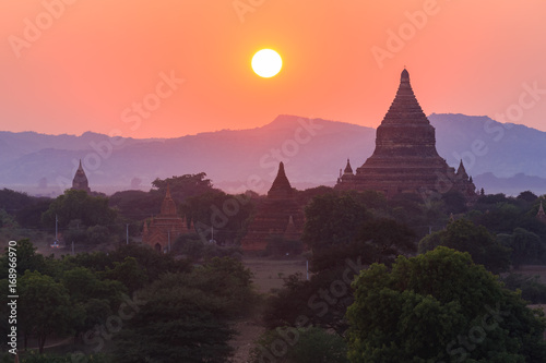 View from afar of the ancient pagodas (stupas) visible among rugged fields and trees of other pagodas and mountains on the horizon during sunset or sunrise, in Bagan, Myanmar (Burma) © CHATCHAI