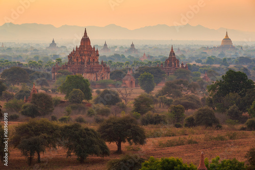 View from afar of the ancient pagodas  stupas  visible among rugged fields and trees of other pagodas and mountains on the horizon during sunset or sunrise  in Bagan  Myanmar  Burma 