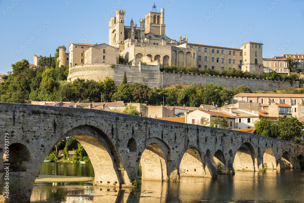 Views at sunset of the French city of Beziers, with trees and the old bridge reflected over the river Orb, and the 13th-century Cathedral of Saint Nazaire in the background
