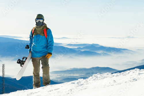 Young snowboarder in helmet standing at the top of a mountain with beautiful sky on background