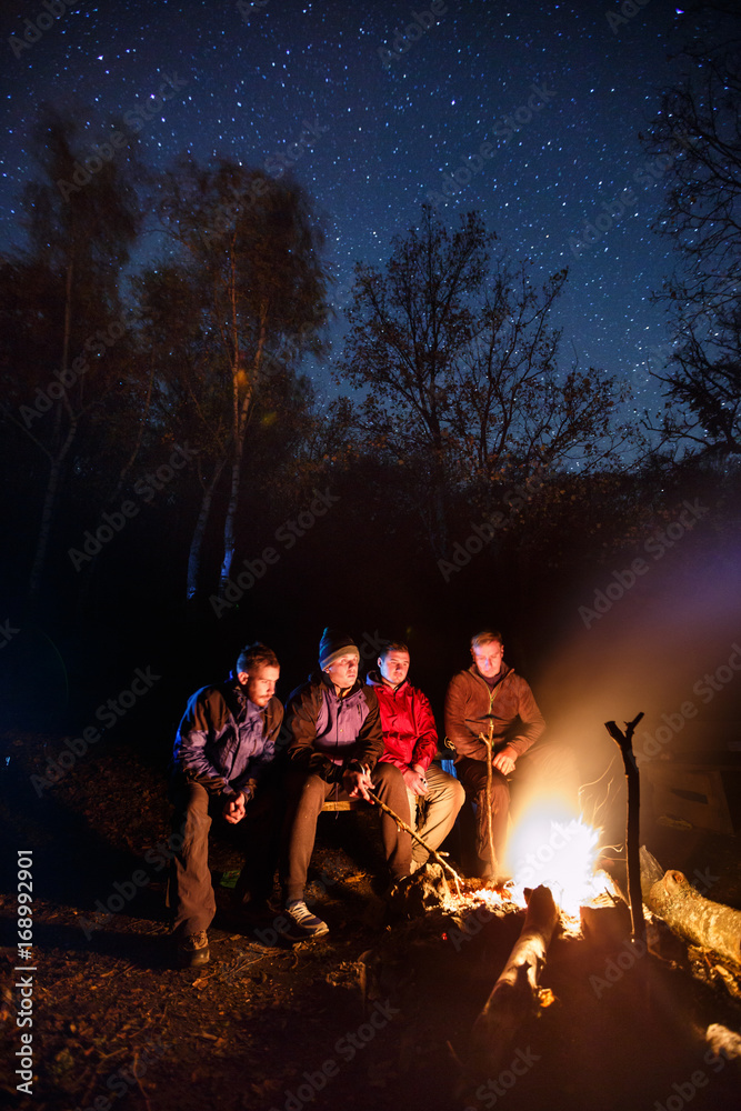 Long exposure photo of four hikers sitting around bonfire and warming at night autumn forest with starry sky on background. Tourists are preparing their dinner on fire.
