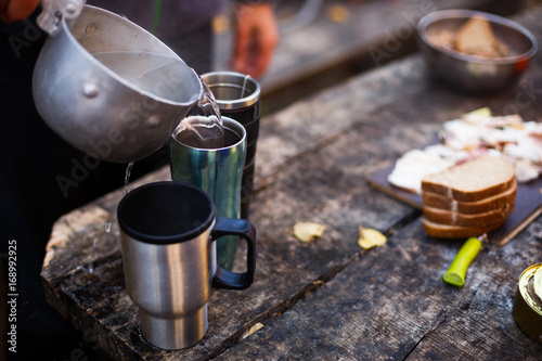 Man pour boiled water to hot mugs on wooden table during breakfast at the forest camp. People on picnic camping at national park and doing lunch tea and coffee. Isolated view