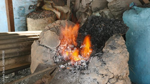 Upper view of a blacksmith's coal forge with a piece of metal heating inside hearth; Blacksmith stirring coal with a fire-stick to keep fire lit