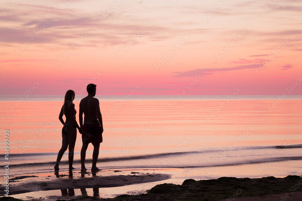 Young couple walking at the seaside in summer evening. Man's and woman's silhouettes on the beach. Sea water reflecting a pink heavenly colors and creating a romantic atmosphere.