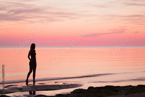 Young woman walking alone at the seaside in summer evening. Woman silhouette on the beach. Sea water reflecting a pink heavenly colors and creating a romantic atmosphere.