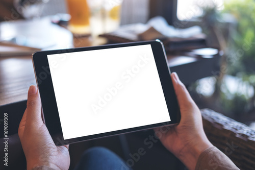 Mockup image of hands holding black tablet pc with white blank screen on wooden table background in cafe