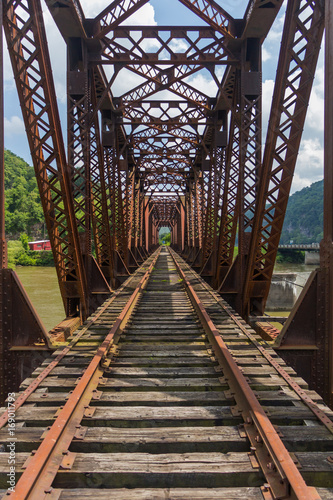 Fotografija A down the track perspective across an old rusted railroad bridge spanning a river with green mountains in the distance with a blue cloudy sky