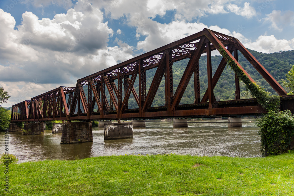 Old rusted railroad bridge crossing a river with green grass, mountains in the distance and a cloudy blue sky overhead. Old rusted railroad bridge crossing a river with green grass, mountains in the distance and a cloudy blue sky overhead.