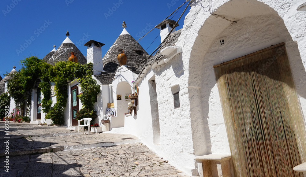 Fototapeta premium ALBEROBELLO, ITALY - JULY 31, 2017: Beautiful street of Alberobello with trulli houses among green plants and flowers, main touristic district, Apulia region, Southern Italy