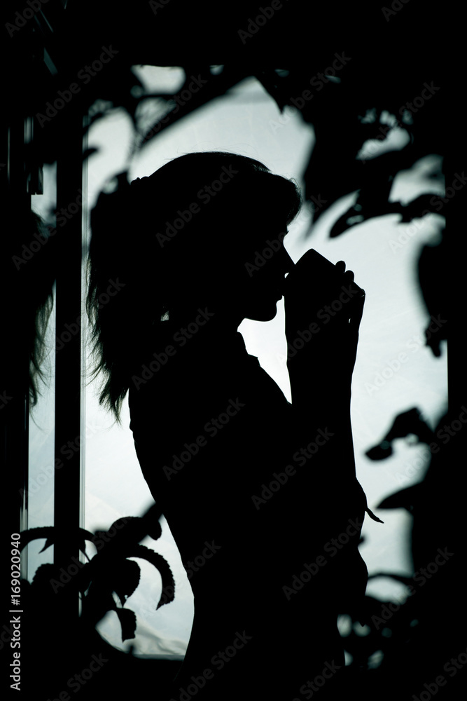silhouette of the figure of a woman drinking hot tea near a window in foliage of a tree